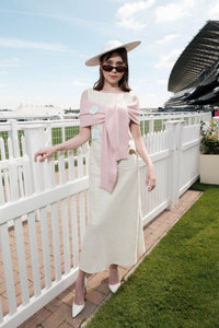 A woman stands outdoors at a racetrack wearing a light cream midi dress with a large pale pink draped wrap detail across the bodice, a matching wide-brim hat, and white pointed heels.